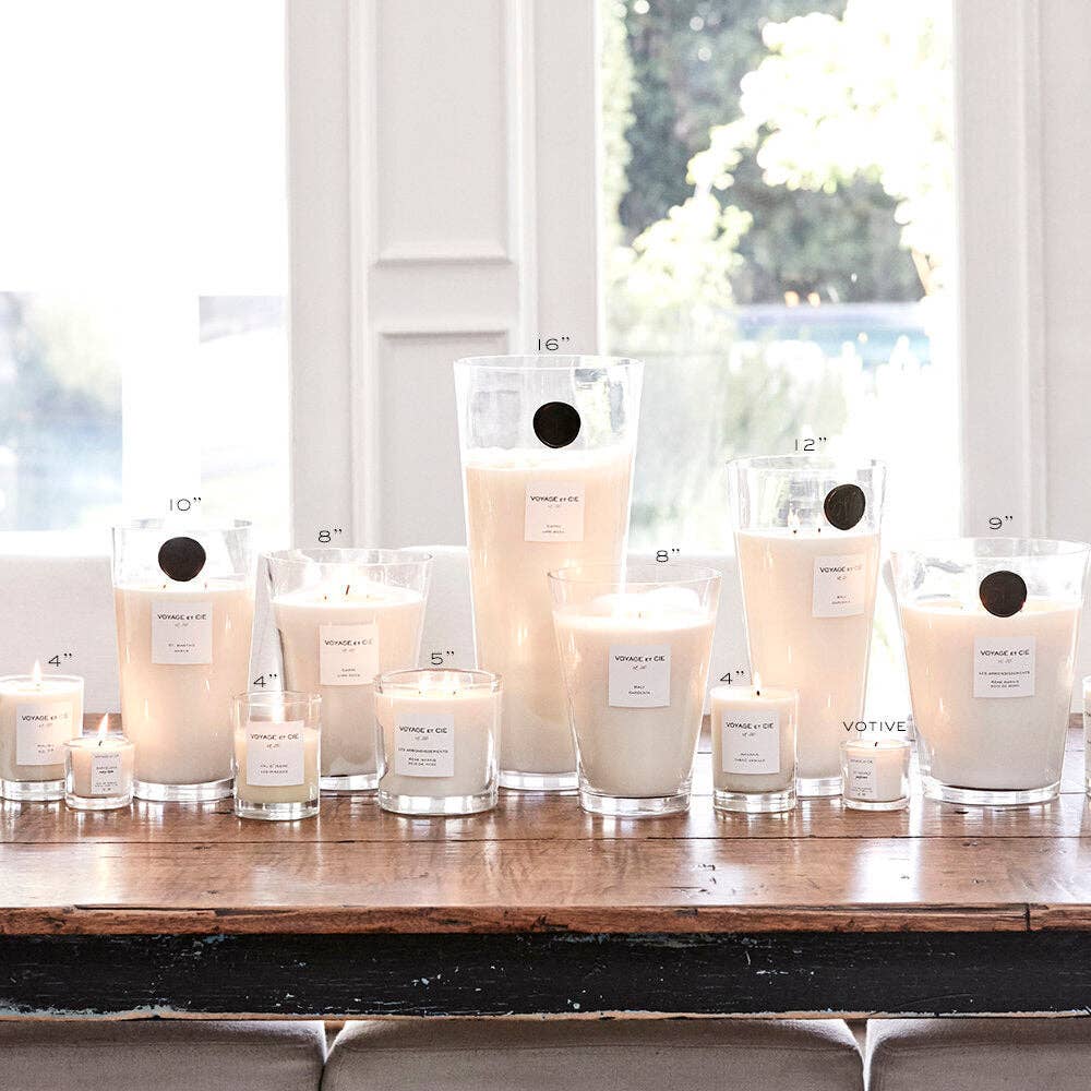 An assortment of white soy candles in various sizes and glass containers, including tall tumblers and smaller jars, arranged on a rustic wooden table in front of a bright window.