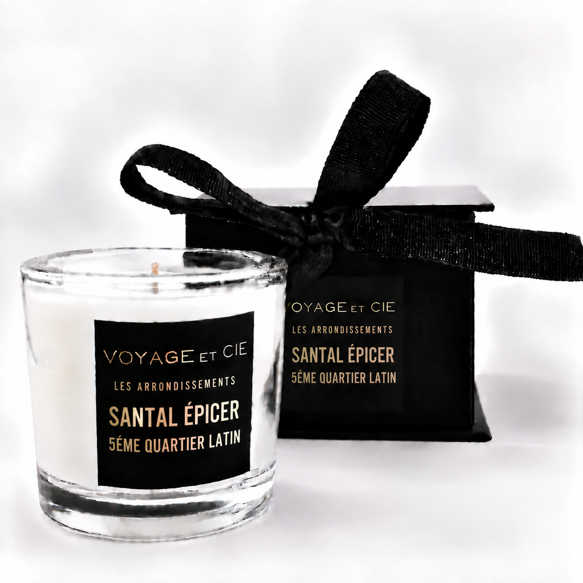 A white soy candle in a clear glass jar with a black label reading 'VOYAGE ET CIE MYKONOS GINGER CITRUS', shown next to its black gift box tied with a black ribbon on a white background.