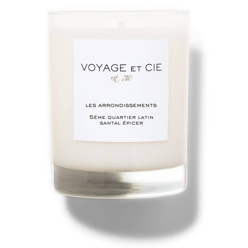 A clear glass jar candle with creamy off-white wax and a white rectangular label reading “VOYAGE ET CIE est. 202°°” and “LES ARRONDISSEMENTS 5ÉME QUARTIER LATIN SANTAL EPICER,” shown on a white background with a soft shadow.