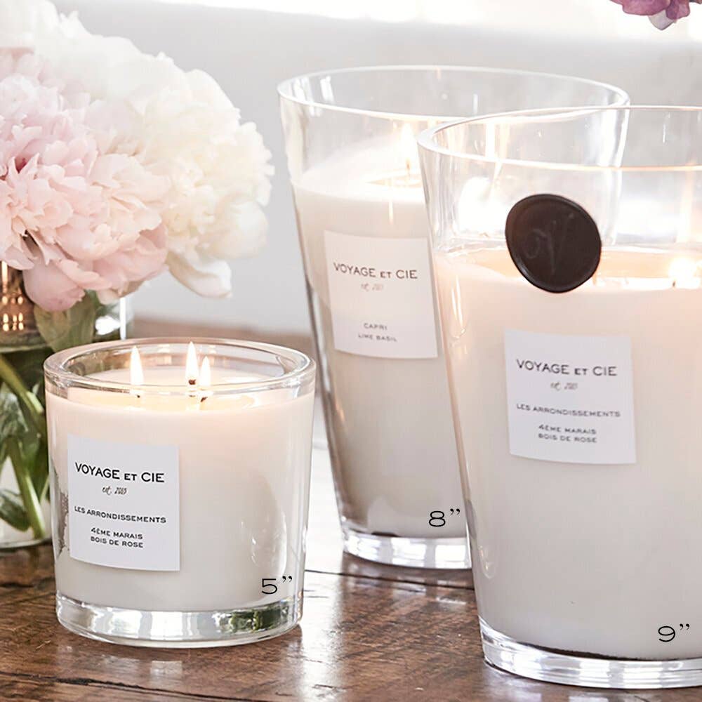 Three lit candles with 'Voyage et Cie' labels on a wooden surface with flowers in the background.