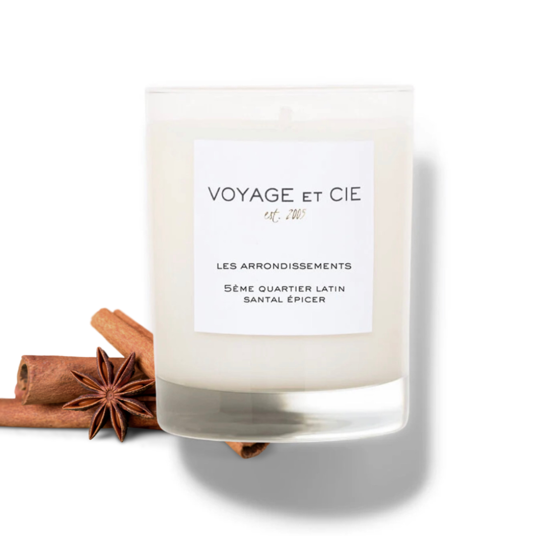 A white soy wax candle in a clear glass jar with a white label reading 'VOYAGE ET CIE', styled with cinnamon sticks and an star anise on a white background.