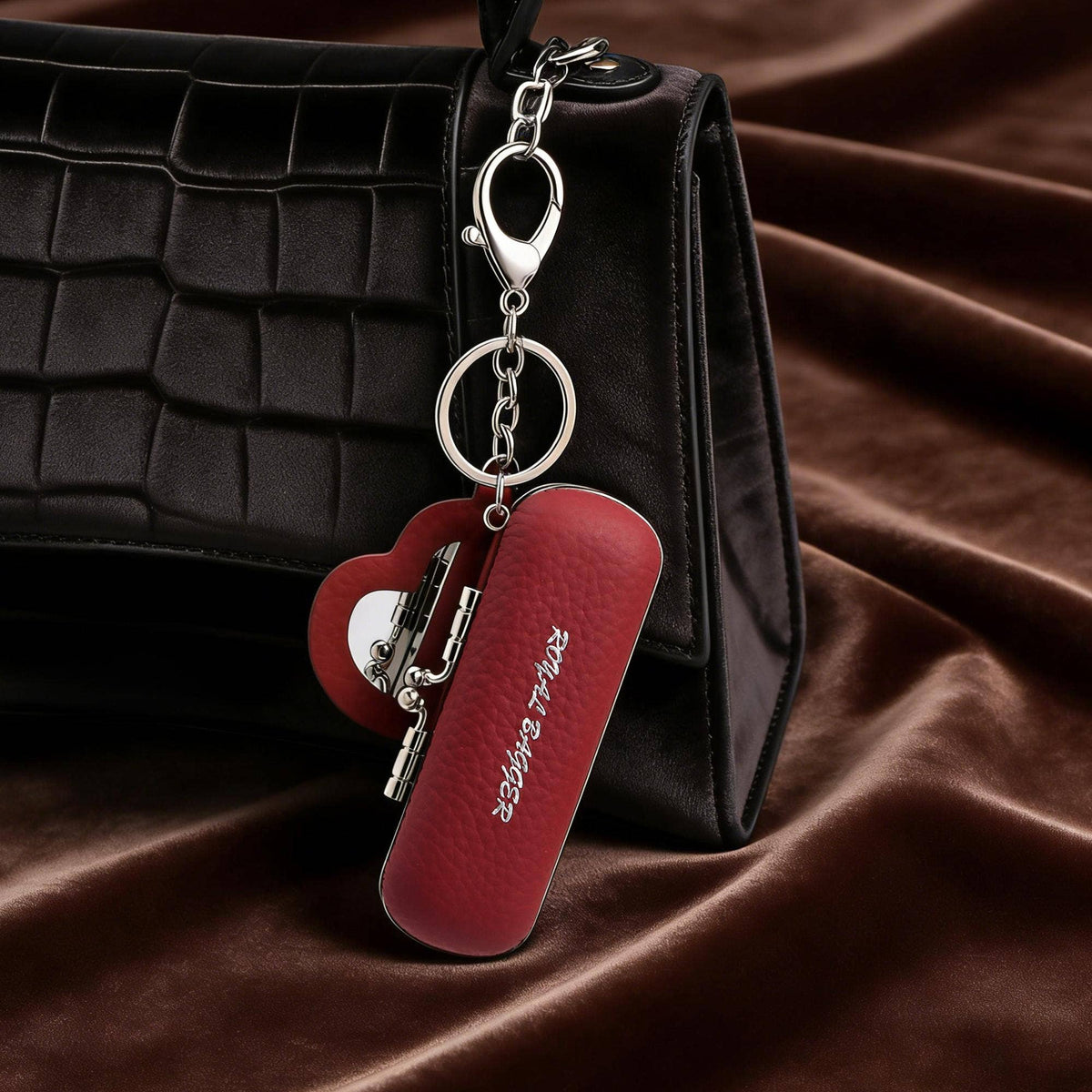 A red textured leather keychain bag with a silver clasp and keyring, shown resting against a black crocodile-embossed handbag on a draped brown satin fabric background.