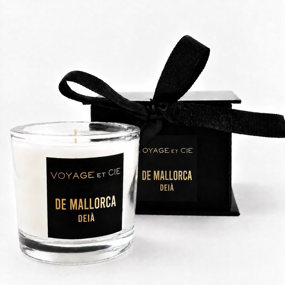 A clear glass jar candle with white wax sits in front of a black gift box tied with a black ribbon. Both pieces feature a black label with gold text reading 'VOYAGE et CIE' and 'ST.