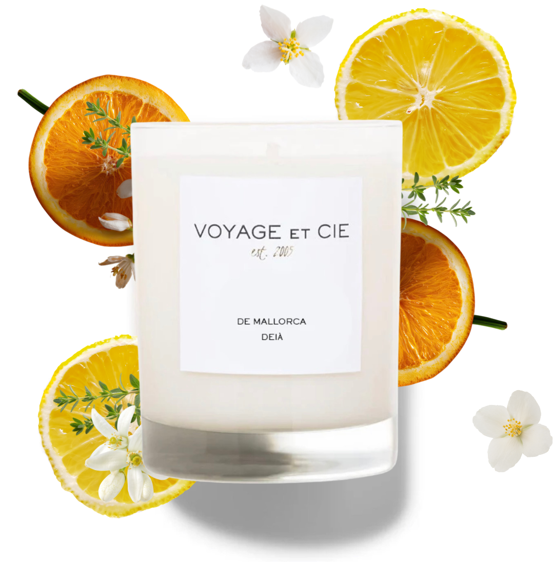 A white scented candle in a clear glass jar with a white label reading 'VOYAGE ET CIE' and 'DE MALLORCA', surrounded by sliced orange, lemon, and small white flowers on a white background.