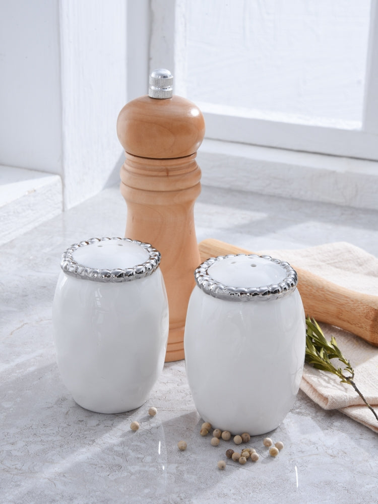 Two white ceramic salt and pepper shakers with silver metal lids, shown next to a wooden pepper mill on a light stone surface.