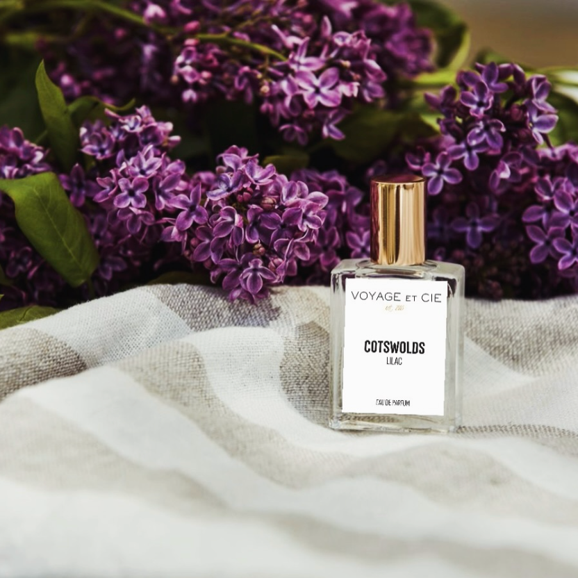 A clear rectangular perfume bottle labeled 'VOYAGE et CIE' and 'COTSWOLDS LUIE' with a gold cap, placed on a white-and-beige checkered cloth in front of purple lilac flowers.