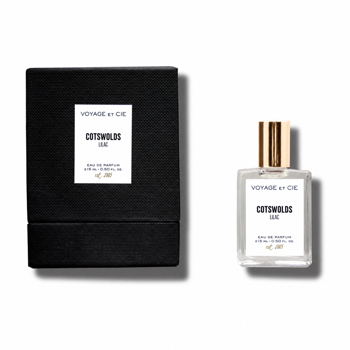 A small clear perfume bottle with a metallic gold cap stands beside its black textured box. Both pieces have white labels reading 'VOYAGE et CIE' and 'COTSWOLDS LEIC'.