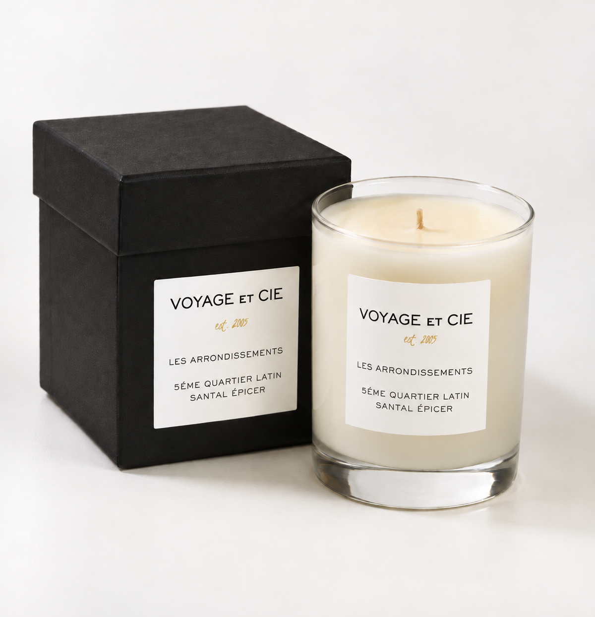 A glass jar candle with cream-colored wax and a white label next to its black gift box, both branded 'VOYAGE ET CIE'.
