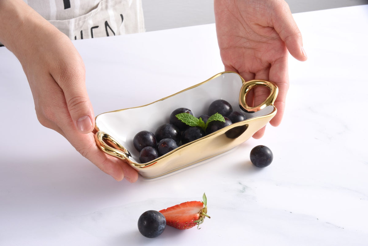 Person holding a white dish with gold rim containing black grapes on a white surface.