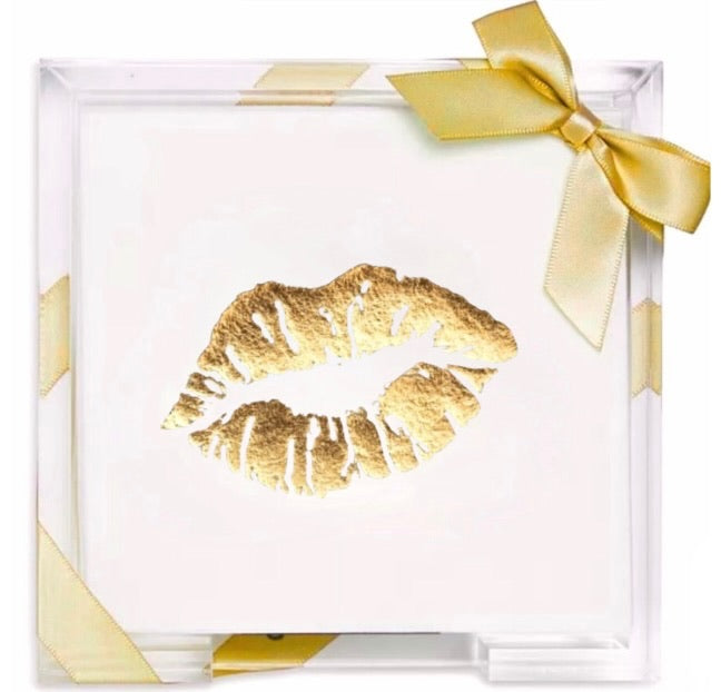 A square clear acrylic gift box containing a white card with a metallic gold lip print in the center, tied with a gold ribbon bow at the top right.