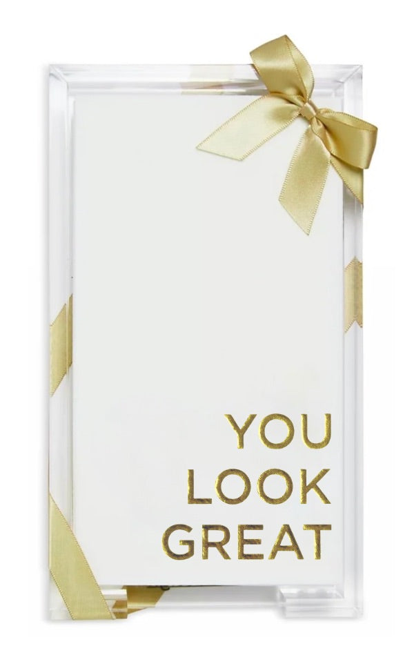 A clear acrylic gift box containing a white card with gold foil text that reads 'YOU LOOK GREAT,' tied with a gold ribbon bow at the top and gold ribbon accents on the sides.