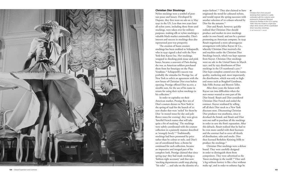 Black and white scene of a woman in a large dress with a hat, sitting at a table with a man, Christian Dior in an elegant room.