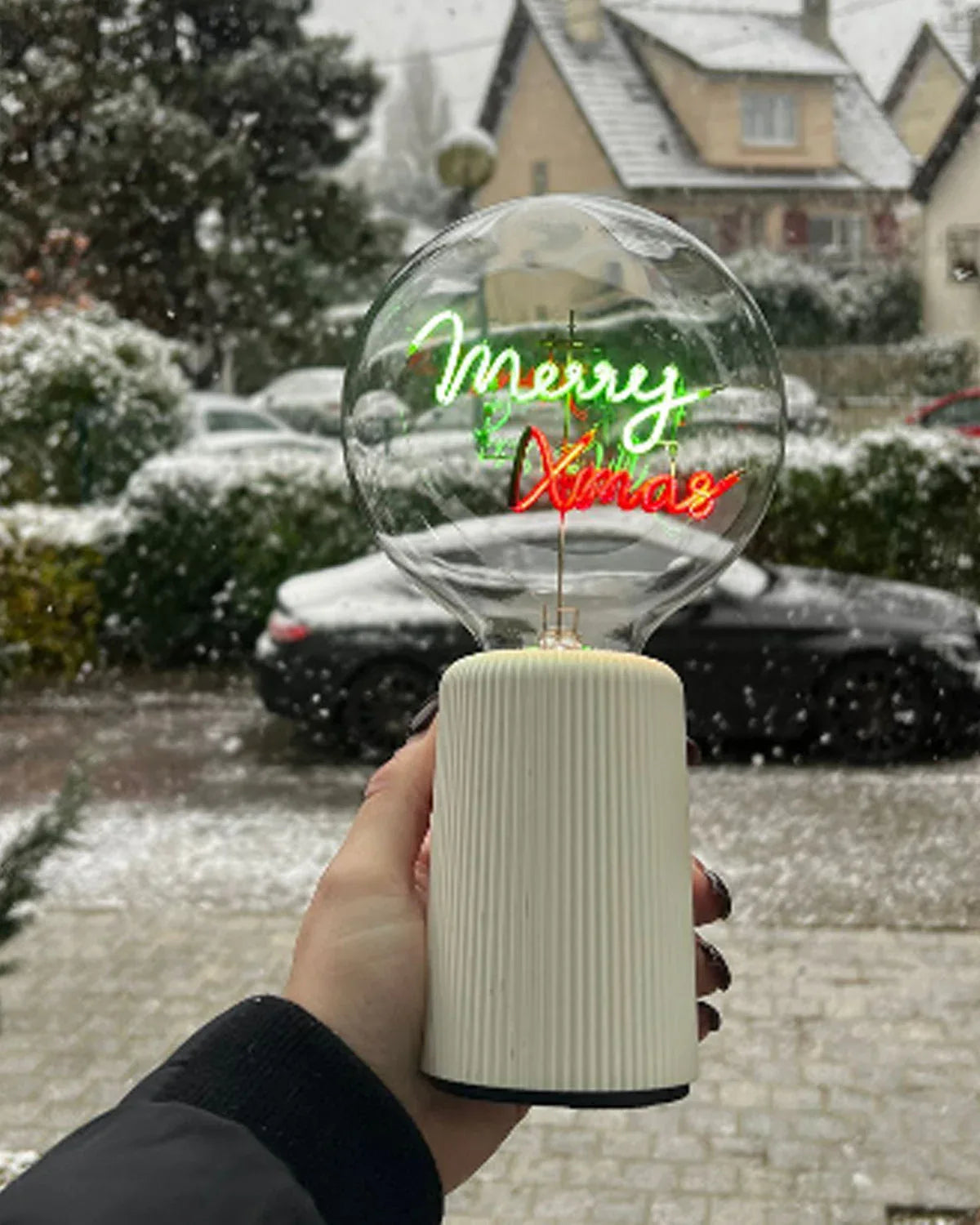 Hand holding a decorative light bulb with 'Merry Xmas' text against a snowy outdoor background.