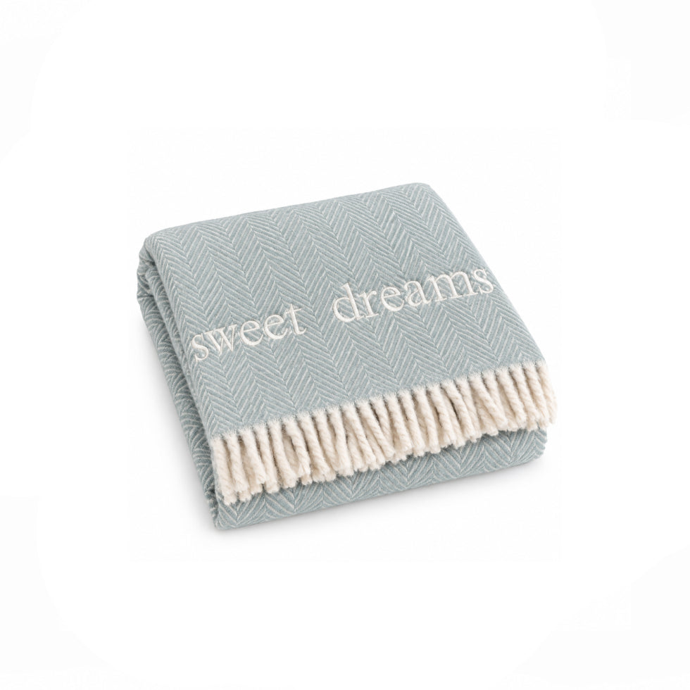 A folded herringbone throw blanket in a dusty sage green color with cream fringe and the words 'sweet dreams' woven in white across the front, shown on a plain white background.