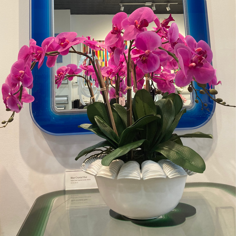 Pink orchid in a white pot on a glass table with a blue-framed mirror in the background.