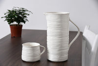 White ceramic pitcher and mug on a wooden table with a plant in the background