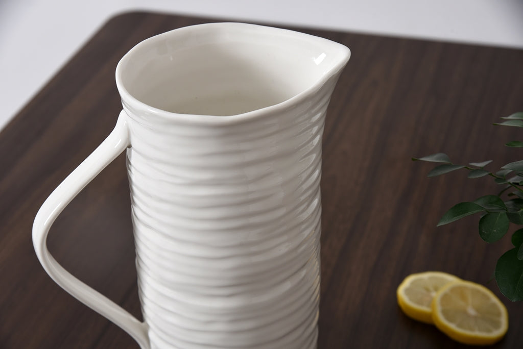 White ceramic pitcher on a wooden surface with lemons and a plant.