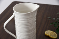 White ceramic pitcher on a wooden surface with lemons and a plant.
