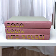 Three pink books with gold text on a white surface Coco, Louis, Life is Gucci