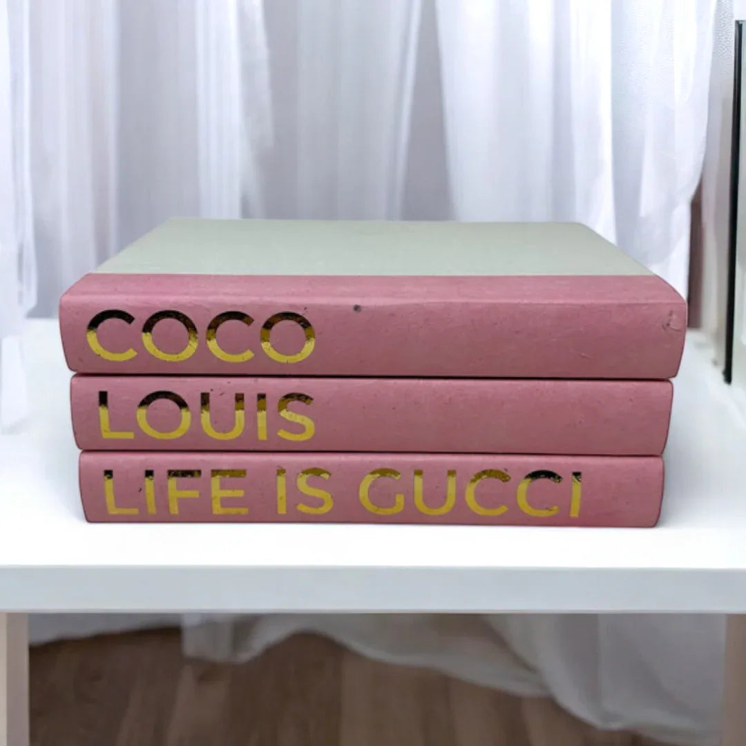 Three pink books with gold text on a white surface Coco, Louis, Life is Gucci