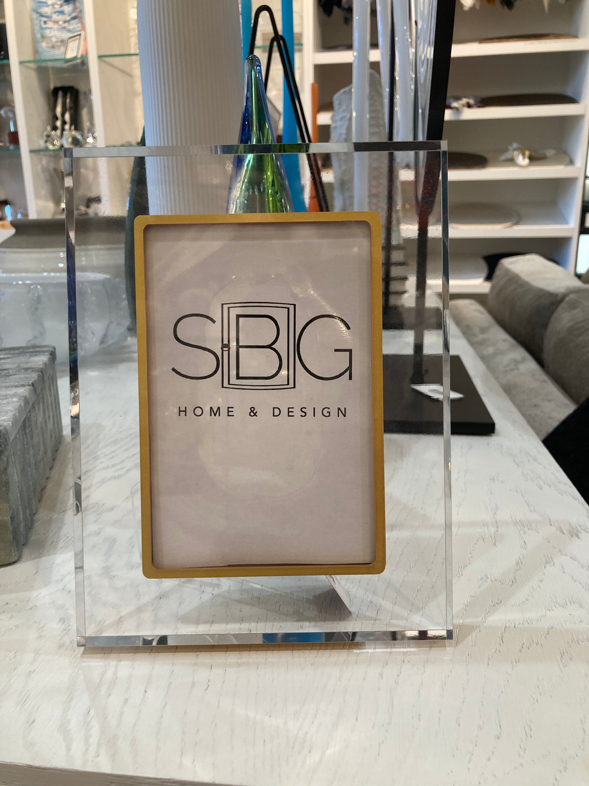 Clear gold acrylic stand with a framed logo on a table, featuring 'SBG Home & Design'.