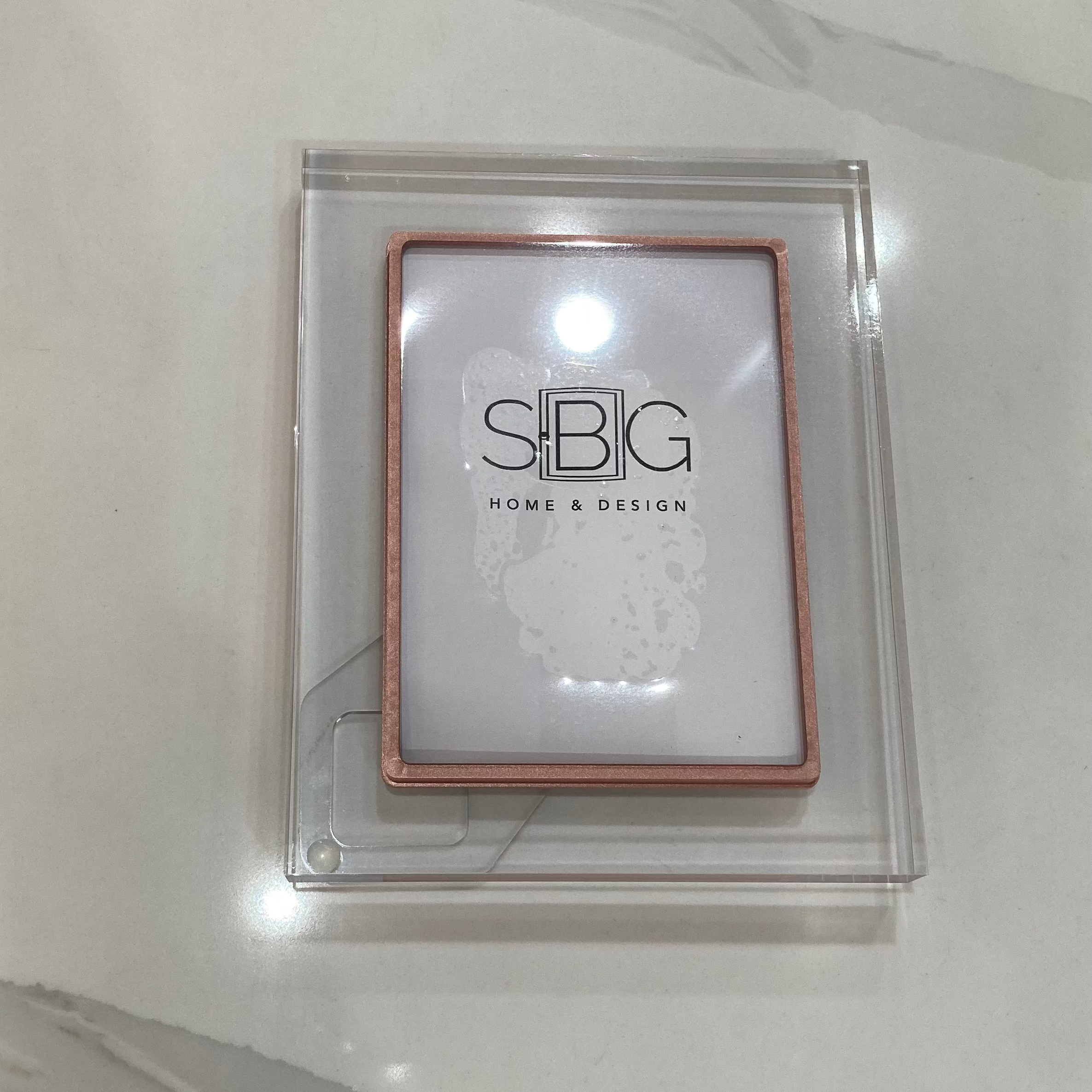 Rose Gold acrylic frame with SBG HOME & DESIGN branding on a marble counter
