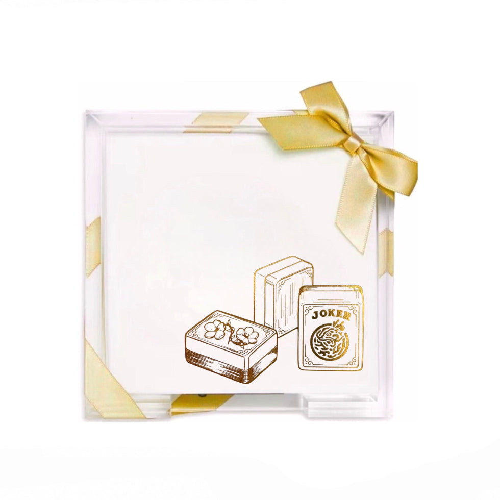 A clear acrylic square box with a white insert, decorated with gold geometric corner accents and a gold ribbon bow. The front features an illustration of three gold playing cards, including a joker card.