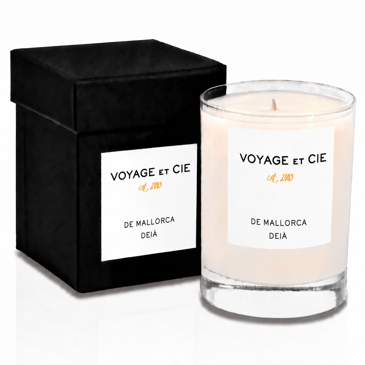 A clear glass jar candle with cream-colored wax sits next to its black presentation box. Both pieces feature a white label with the brand name 'VOYAGE ET CIE' and the scent 'ST.