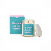 A lit candle in a clear glass jar with a turquoise label reading 'Besties for the resties' in pink text, shown next to its matching turquoise box with a pink top and a white lid on a white background.
