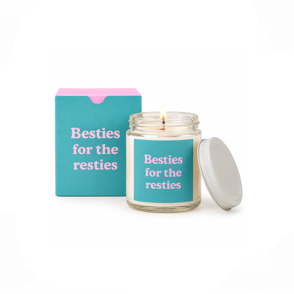 A lit candle in a clear glass jar with a turquoise label reading 'Besties for the resties' in pink text, shown next to its matching turquoise box with a pink top and a white lid on a white background.