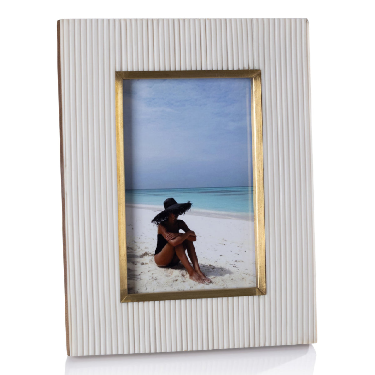 A vertical rectangular picture frame with a ribbed off-white textured border and a thin gold inner trim, holding a beach photograph of a woman sitting on the sand.