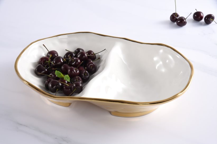 An asymmetrical white ceramic serving bowl with a gold rim, shown on a white marble surface and holding a cluster of dark cherries.