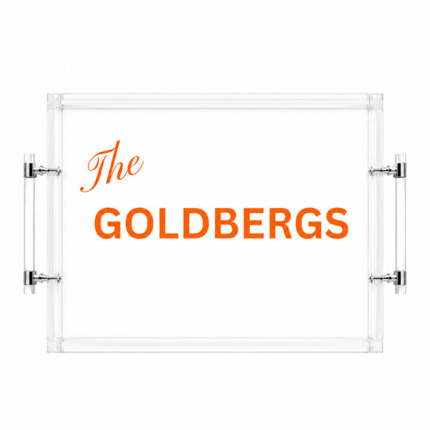 A clear acrylic serving tray with a white insert, featuring the text 'The GOLDBERGS' in orange. The tray has two vertical clear handles with silver metal brackets on the left and right sides, shown against a plain white background.