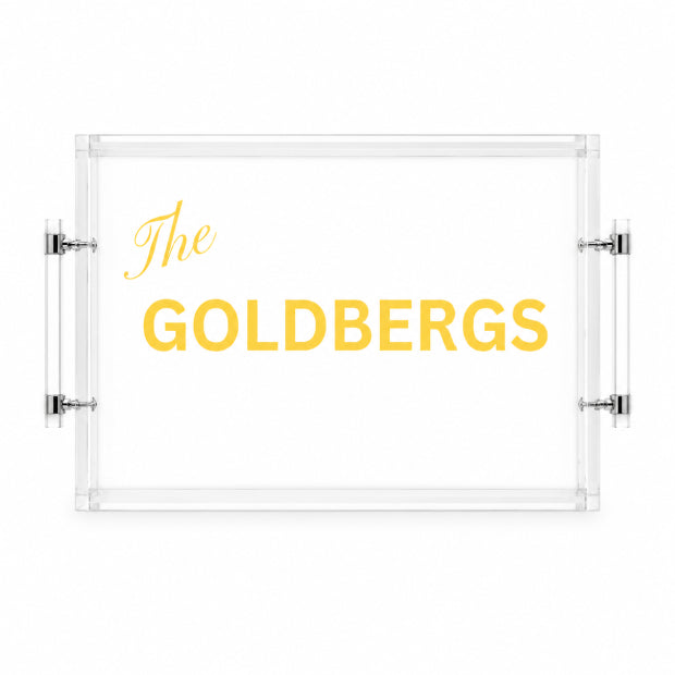 A clear acrylic serving tray with silver metal handles, featuring the text 'The GOLDBERGS' printed in yellow on a white background.