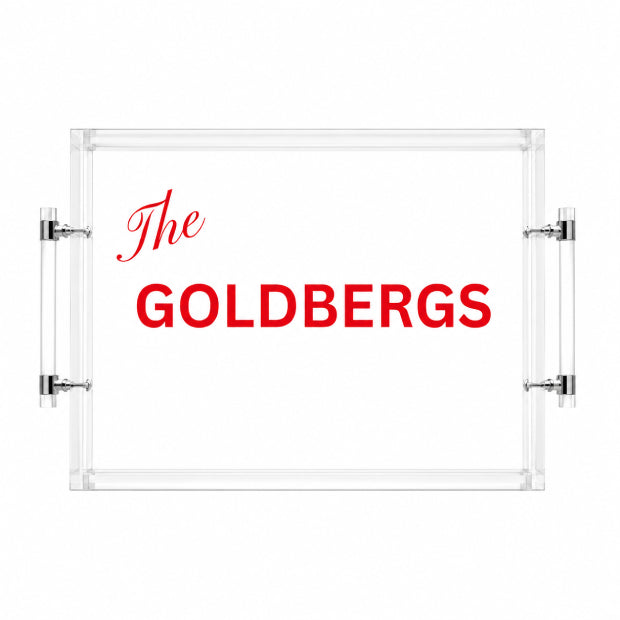 A clear acrylic serving tray with two vertical glass handles and silver metal brackets, featuring the red text 'The GOLDBERGS' printed on a white background.
