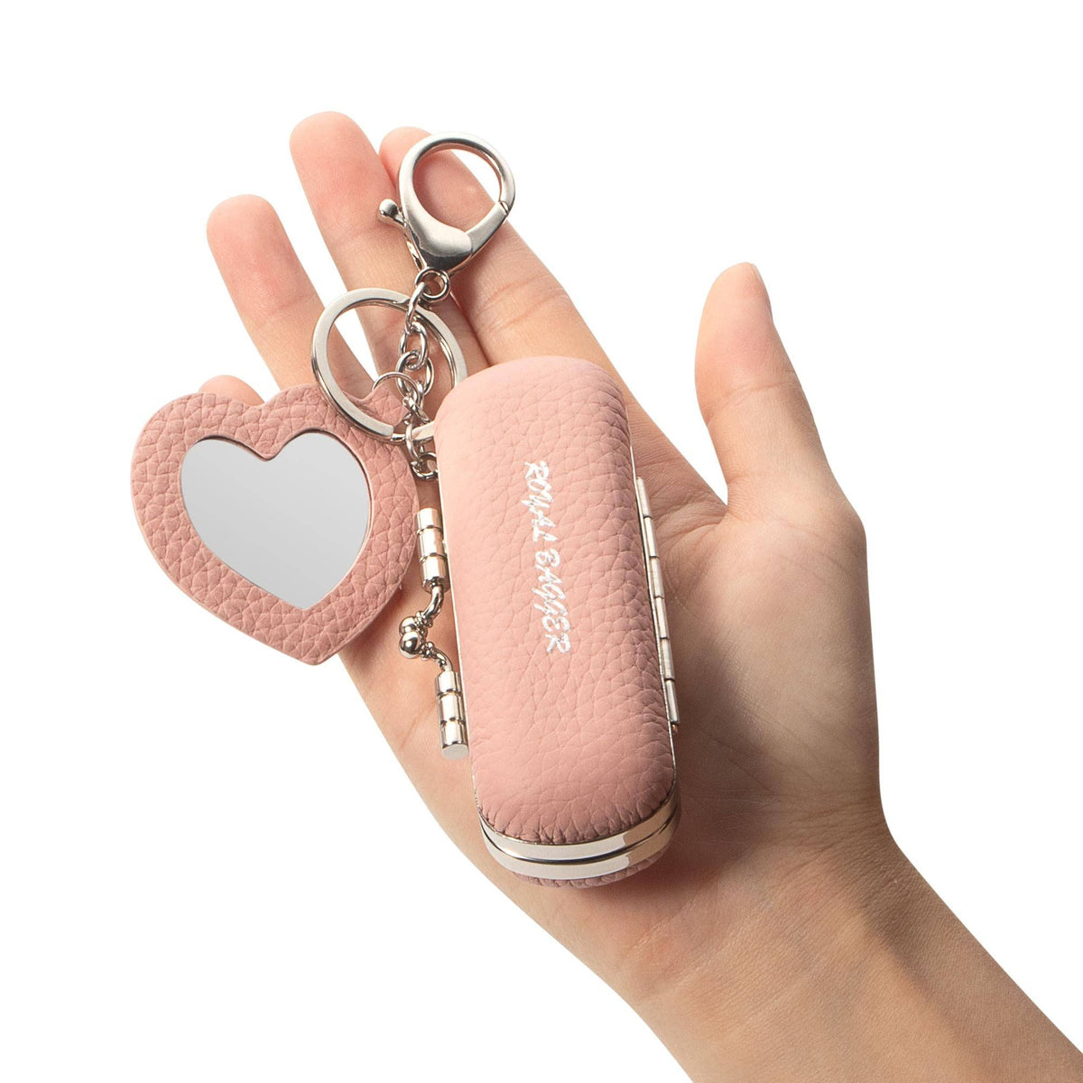 A hand holding a pink pebbled leather keychain compact mirror with a heart-shaped mirror and silver metal keyring on a white background.
