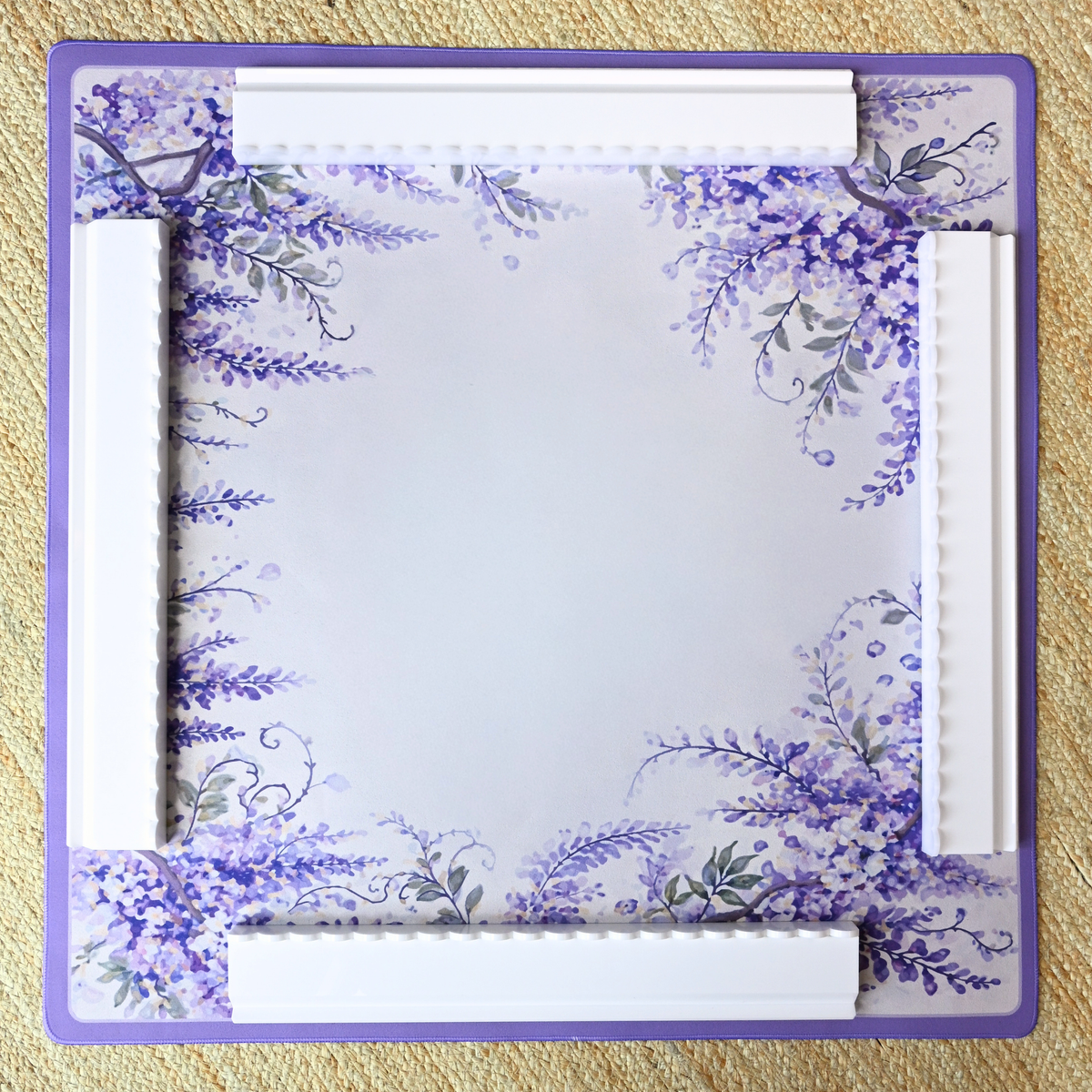 A square mahjong mat with a lavender and purple floral border design, featuring a large blank center and white embossed rectangular accents along the edges, photographed flat on a textured beige surface.
