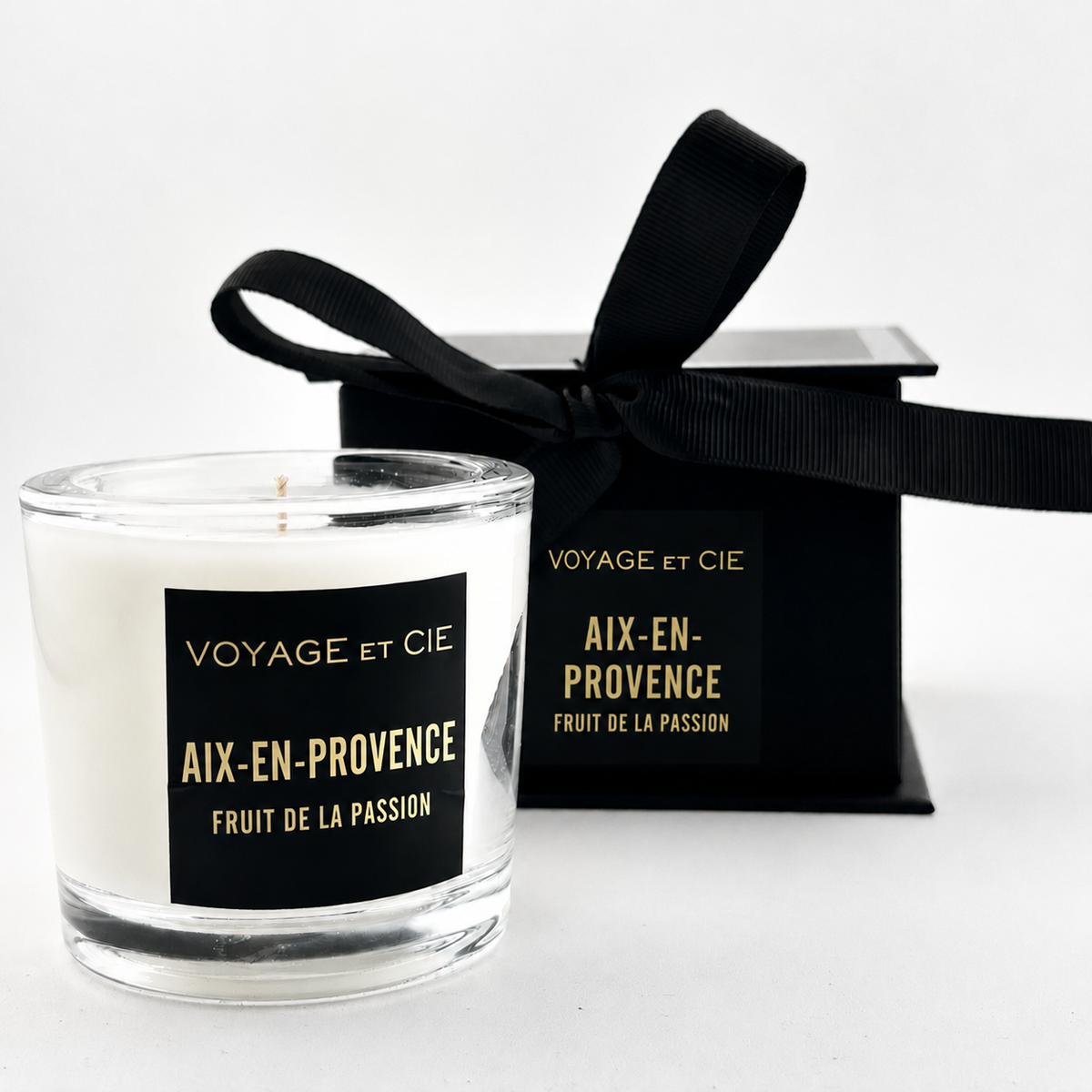 A white soy candle in a clear glass jar with a black label reading 'VOYAGE et CIE TULUM COCONOIX', shown in front of its black gift box tied with a black ribbon. The box has a matching label.