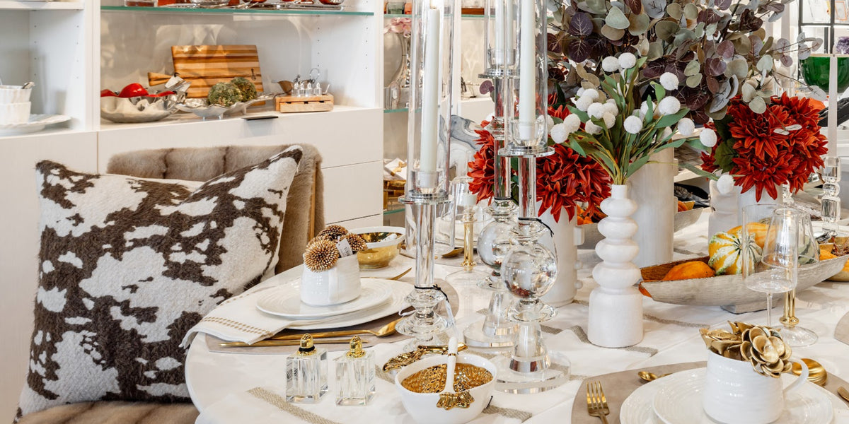 A styled dining table in a bright interior, featuring white plates, clear glassware, gold-toned cutlery, white candlesticks, and floral arrangements with red and white flowers. A white chair with a brown-and-white cowhide-pattern pillow is visible on the left.