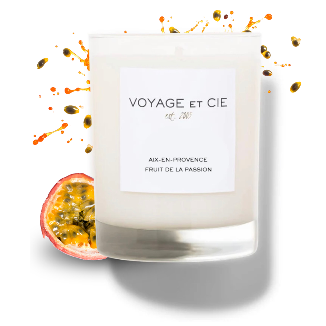 A white soy candle in a clear glass jar with a white label reading 'VOYAGE ET CIE' and 'AIX-EN-PROVENCE FRUIT DE LA PASSION.' A sliced passion fruit sits beside the jar, with orange and yellow passion fruit splatters scattered around on a white background.
