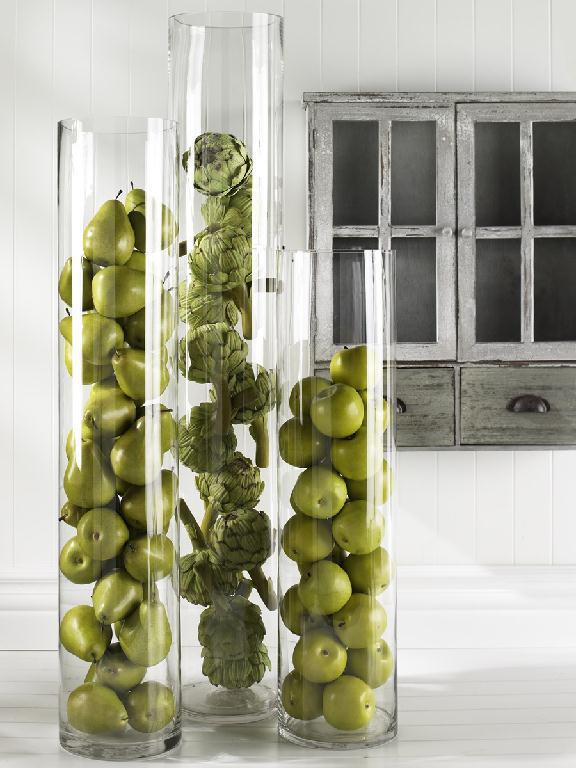 Three tall glass vases filled with green apples, pears and artichoke and leaves on a white surface.