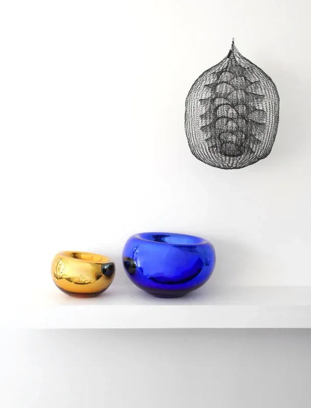 Two glass vases, one gold and one blue, on a white surface with a black wire sculpture in the background.
