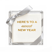 A square clear acrylic gift box containing white paper napkins with gold foil text reading 'HERE'S TO A sweet NEW YEAR,' tied with a silver satin bow on a white background.