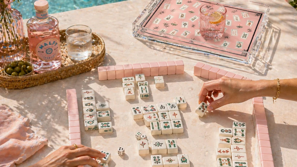 A styled game scene around a pool table showing a pink Mahjong-style tile game in progress. Two hands are visible arranging white and pink tiles on a light stone surface, with a pink tray, glass, bottle, and olives nearby.