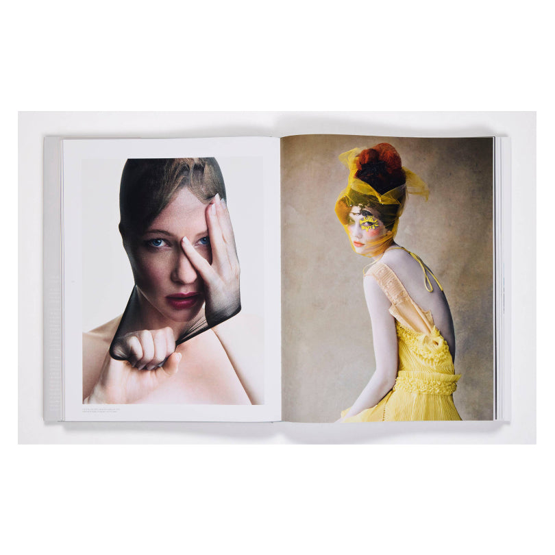An open art book or magazine displayed against a white background, showing two fashion portrait pages side by side. The left page features a close-up portrait of a woman with her hand over her face, and the right page shows a woman in profile wearing a bright yellow dress and headpiece against a soft beige background.
