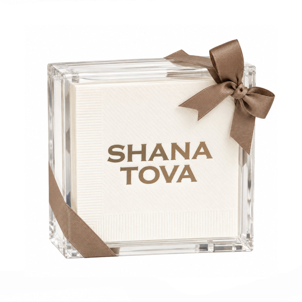 A clear acrylic square box containing white textured paper napkins, tied with a brown ribbon bow and ribbon accent, with 'SHANA TOVA' printed in brown on the front, on a white background.