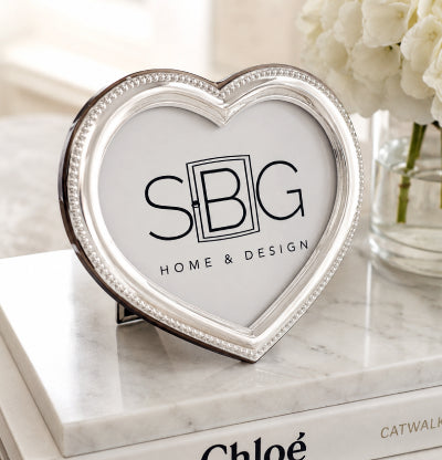 A heart-shaped silver picture frame with a beaded border and a white insert displaying the 'SBG HOME & DESIGN' logo, styled on a white marble book with white flowers in the background.