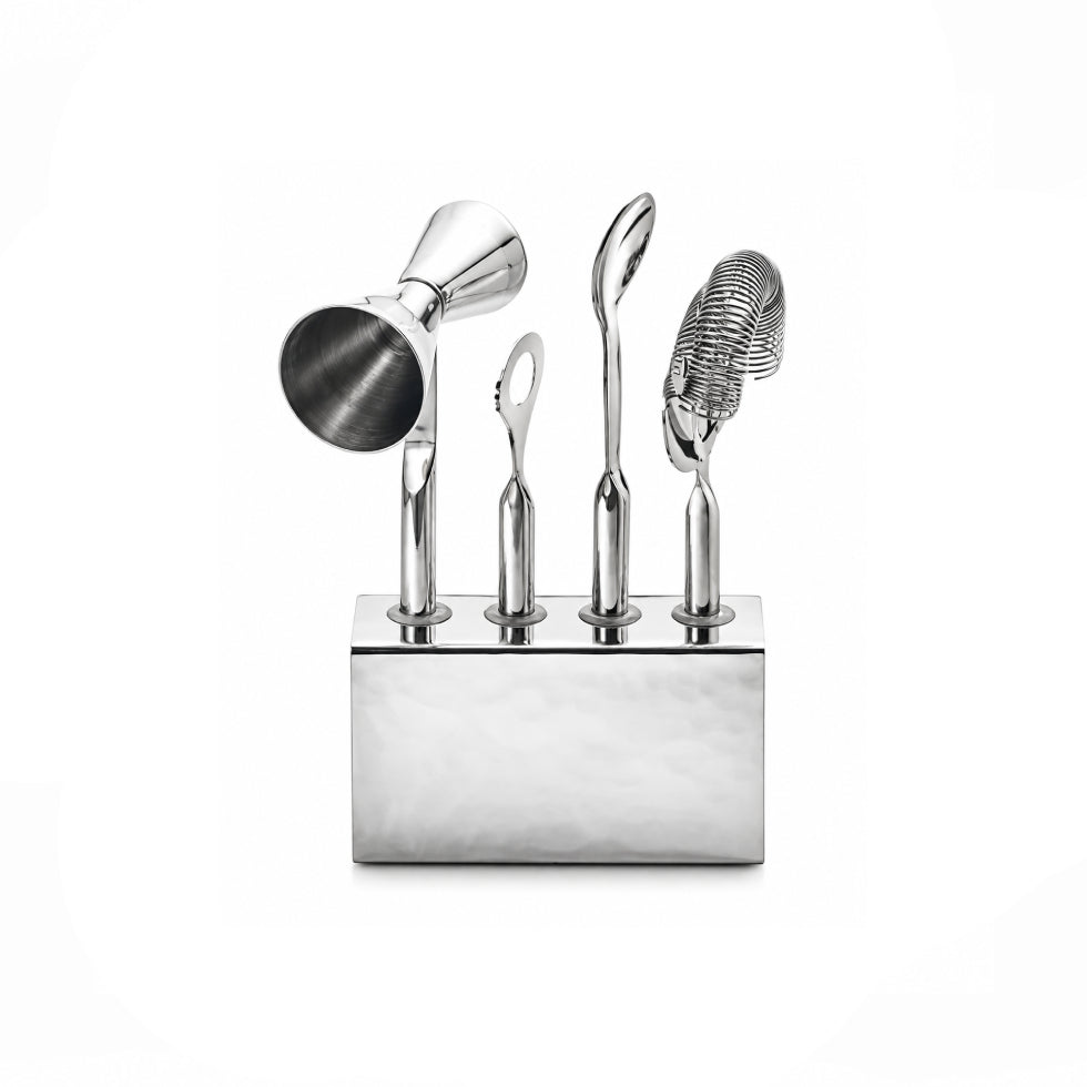 A polished stainless steel cocktail tool set with a rectangular base holding five tools: a jigger, a spoon, a strainer, a small loop tool, and a bar spoon, shown on a white background.